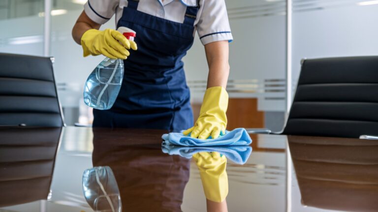 Büro-Reinigungspersonal reinigt einen Konferenztisch. Eine engagierte Reinigungskraft in blauer Uniform und gelben Handschuhen reinigt akribisch einen Glastisch in einem modernen Bürogebäude.